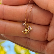 Load image into Gallery viewer, A close-up of a delicate gold chain necklace with three charms, including a small evil eye charm and a rectangular pink sapphire charm, held in a person's hand against a blurred outdoor background.
